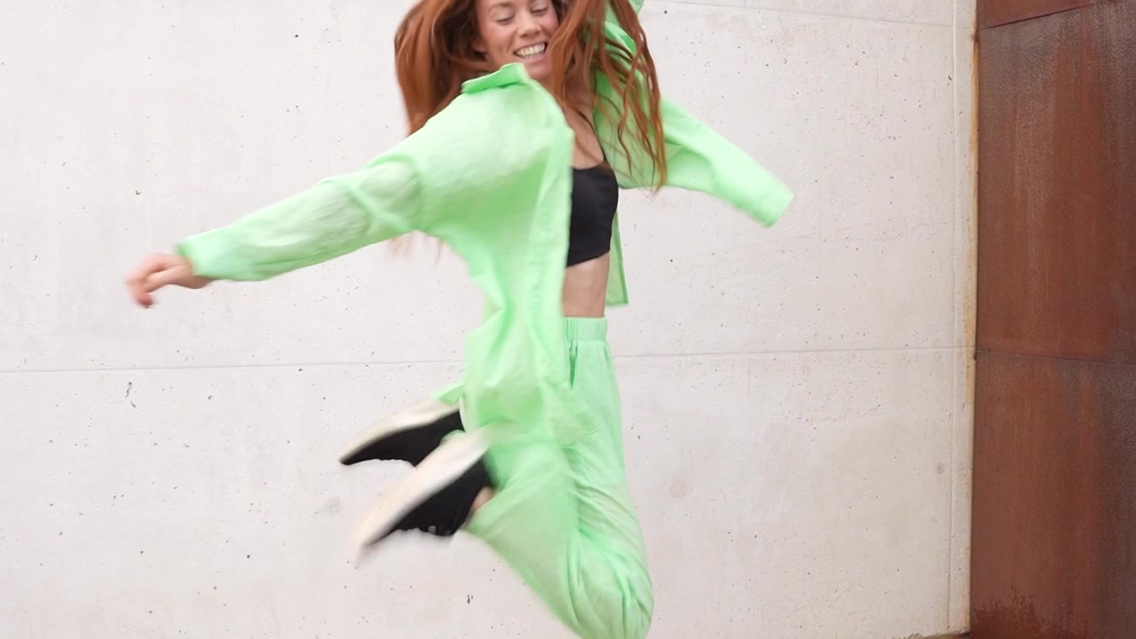 Una donna gioiosa che balla energicamente con un abito verde su sfondo di una parete bianca.