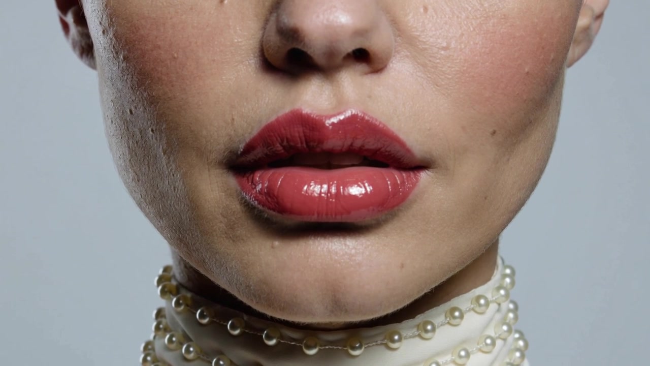 Primo piano del volto inferiore di una modella truccata in studio, con la mascella che si muove, le labbra che si dischiudono, rossetto rosso e una collana di perle.