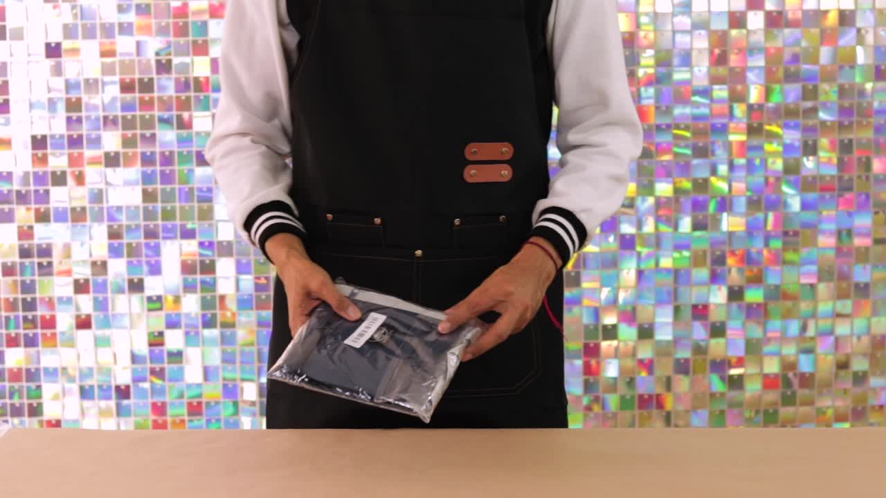 Person wearing an apron unpacks small item from plastic packaging in front of colorful backdrop