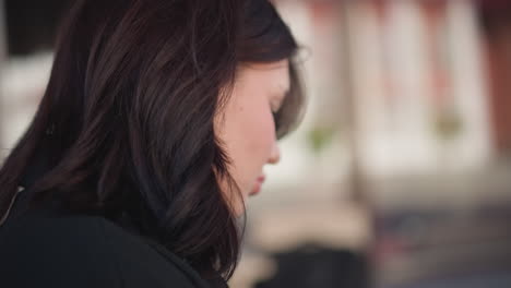 side profile of a young woman with dark brown hair in soft focus, lost in thought, with blurred architectural structures in the background