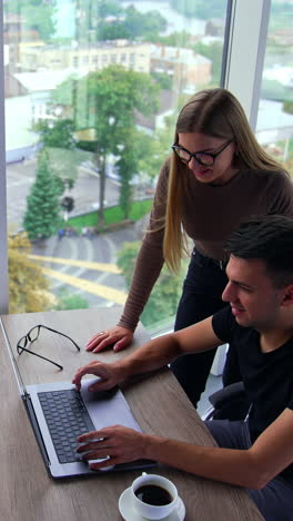 young people working in the office. colleagues look at the screen talking and smiling. beautiful city view in the panoramic windows. high angle perspective. vertical video
