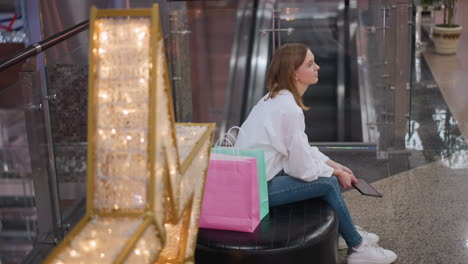 young woman seated on bench in shopping mall holding tablet surrounded by festive decorations, illuminated star decor, and shopping bags, with escalators in background