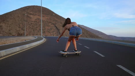 Die-Zeitlupenaufnahme-Zeigt-Eine-Frau-Beim-Skateboarden-Auf-Einer-Straße-Bei-Sonnenuntergang,-Mit-Bergen-Und-Einem-Faszinierenden-Himmel-Als-Hintergrund.-Sie-Ist-In-Shorts-Gekleidet