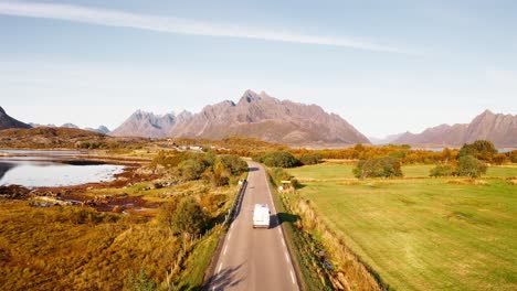 Aerial-view,-rising-up-above-a-road-with-a-white-campervan-making-its-way-towards-the-gorgeous-mountains-in-the-background,-blue-sky-day-of-Fall,-Lofoten-Islands,-Norway