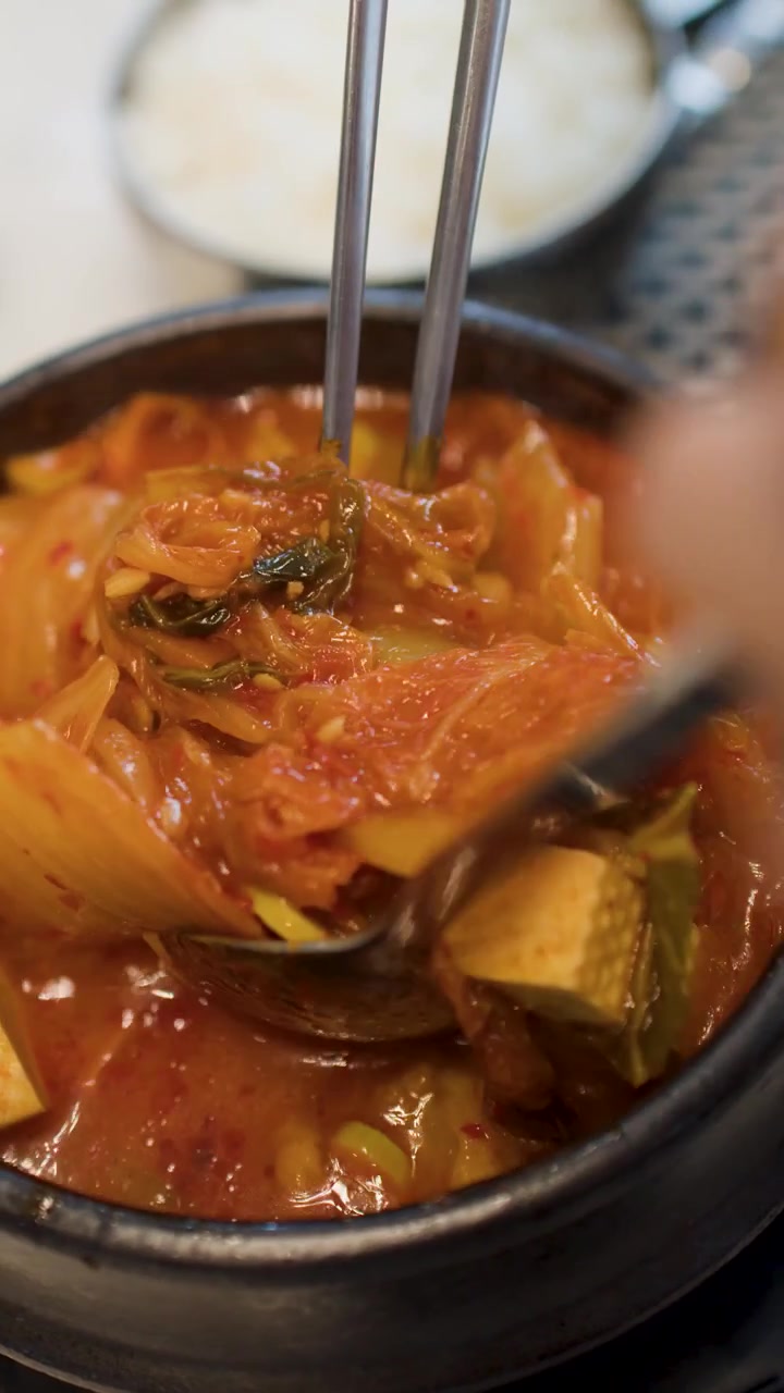 Rekaman jarak dekat semangkuk kimchi jjigae yang mengepul sedang diaduk dengan peralatan logam tradisional