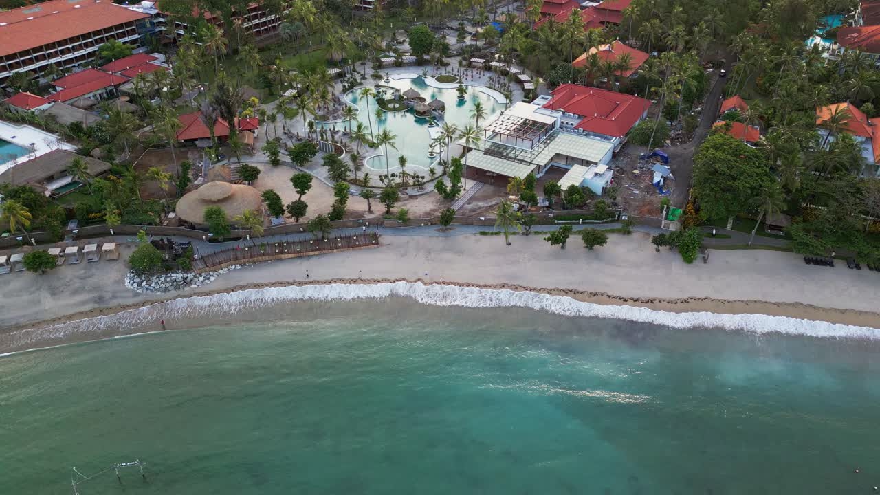 Cinematic drone view of tropical Bali beach resort with palm trees swimming pool and calm blue lagoon beside a wide sandy shoreline with gentle wave movement during golden hour