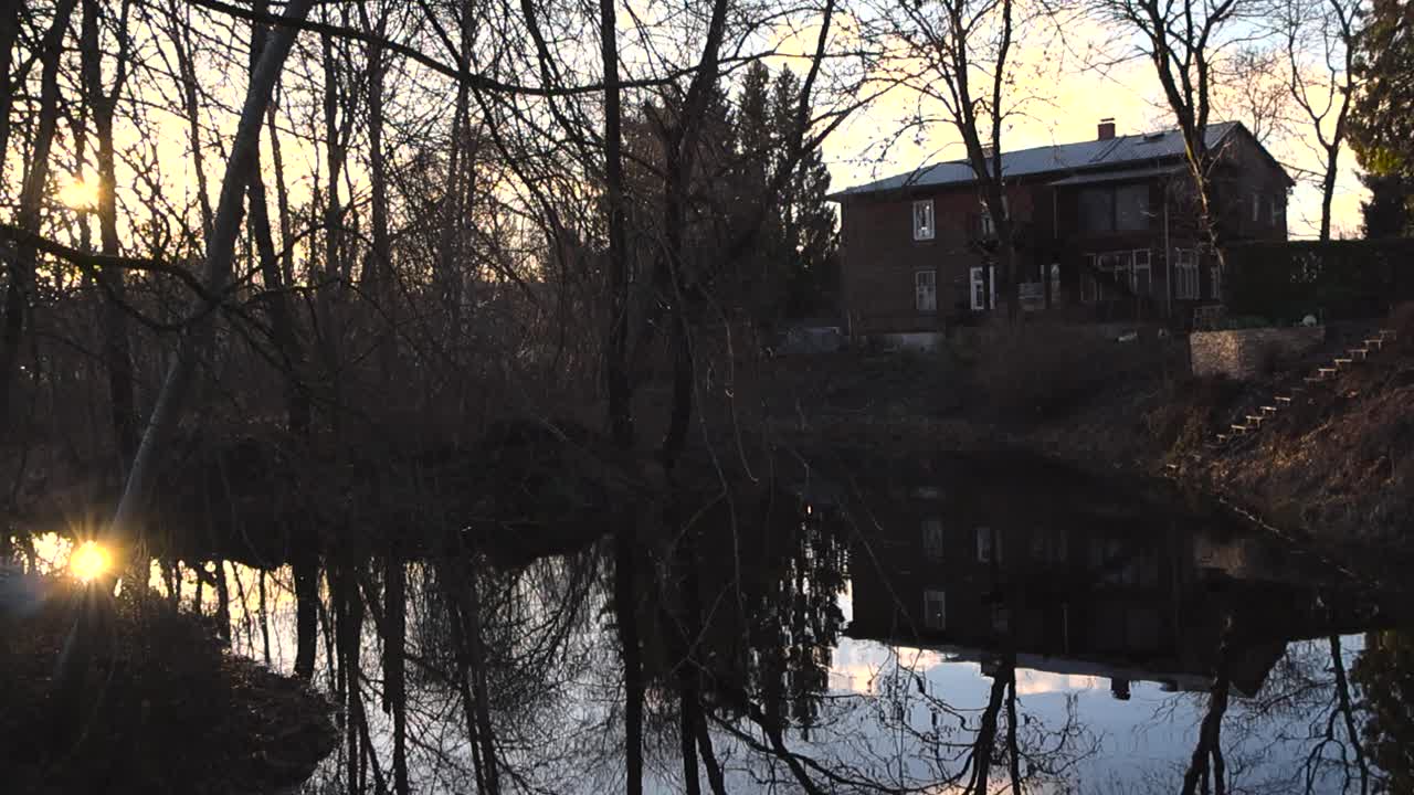 Slowly panning footage of a dark reflective river water in autumn nature during sunset or sunrise with sun rays, lens flare. River banks are dark and have a house on it that is reflecting in water.