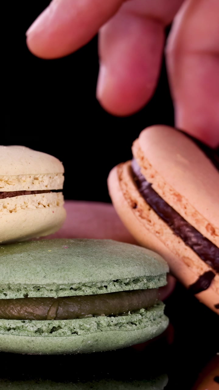 A human hand delicately places assorted pastel macarons in a neat row on a glossy black surface, under bright studio lighting with a close-up perspective
