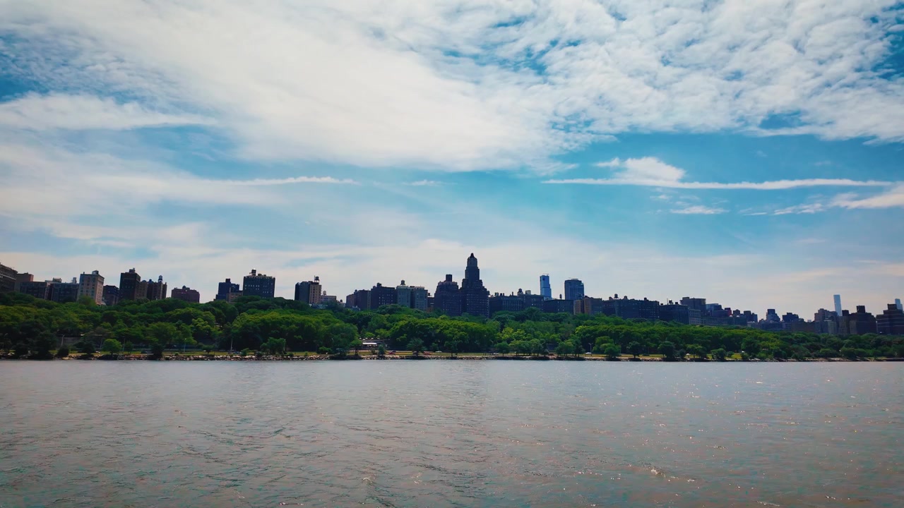 Barco navegando pelo Rio Hudson, passando pelo Upper West Side e Riverside Park. Ampla vista panorâmica do horizonte de Manhattan com vegetação exuberante e céus azuis de verão.