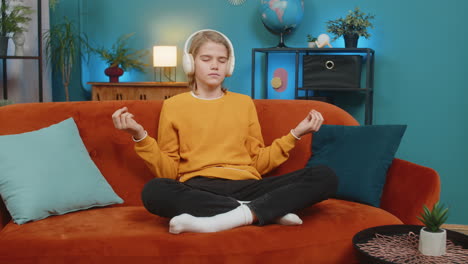 teenager boy relaxing on home sofa meditating in lotus pose with headphones after long school day