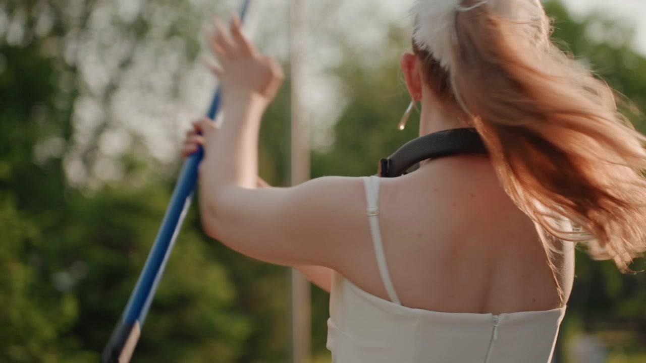 Una giovane donna si allena nel salto con l'asta all'aperto, tenendo un'asta blu in varie posizioni. La donna indossa cuffie al collo e una canotta sportiva su uno sfondo di parco verde con alberi.