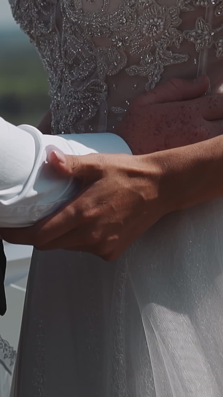 Newlywed groom holds waist of young wife in white fluffy dress with lacy top on riverbank at wedding closeup slow motion. Happy couple at marriage day
