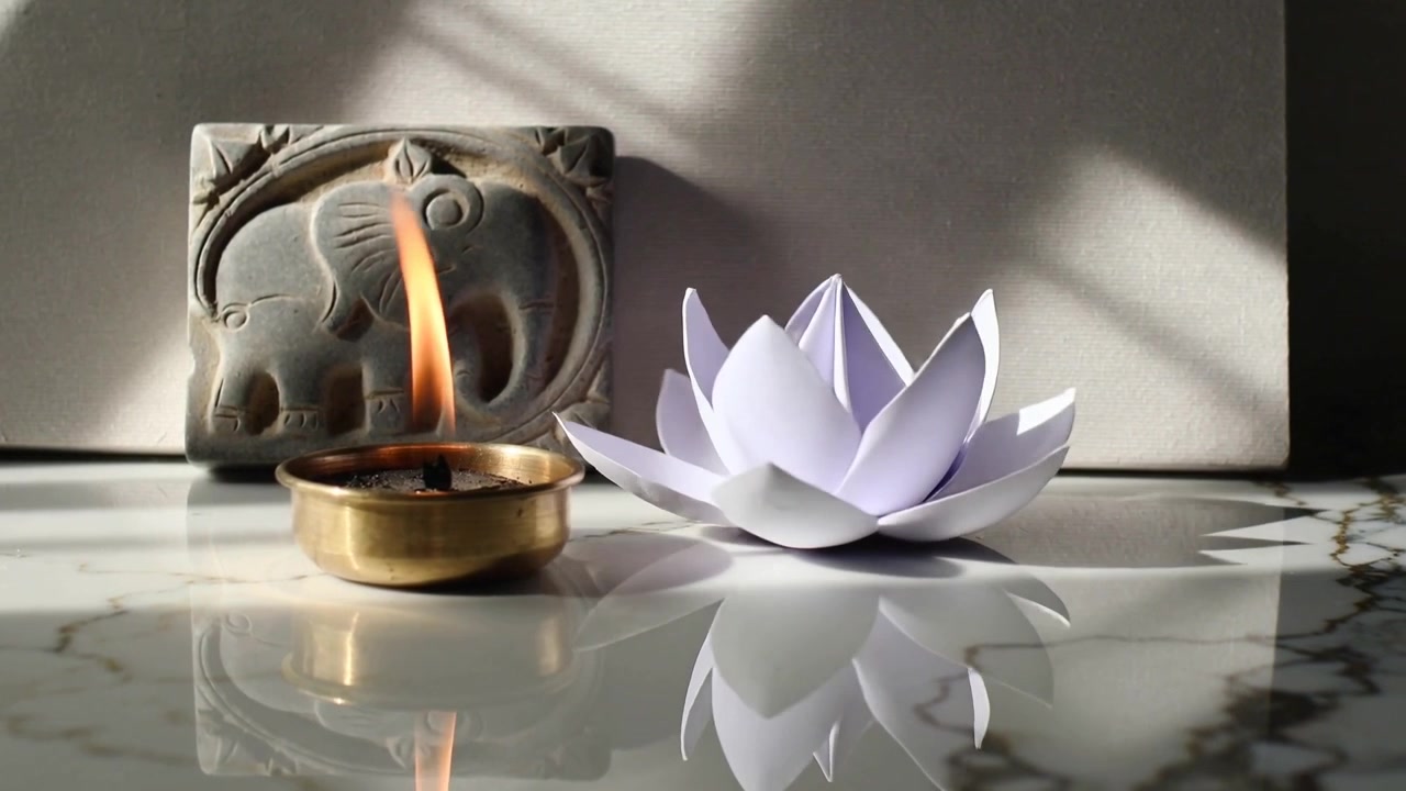 Un raggio di sole che si sposta, spingendo una diya di ottone tremolante e un loto di carta che si inclina su un piano di marmo e una piastrella di pietra.