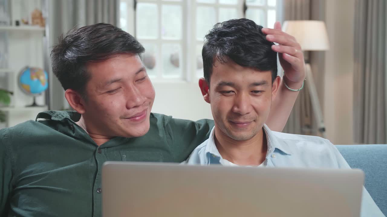 Asian Gentle Gay Couple Using Laptop Computer, While Sitting On A Couch In Cozy Stylish Apartment. Adult Boyfriends Online Shopping On Internet, Watching Funny Videos On Streaming Service