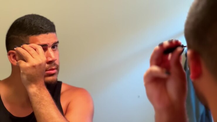 Gay mixed race man looking into mirror adding eyebrow makeup in bathroom, close up LGBTQ