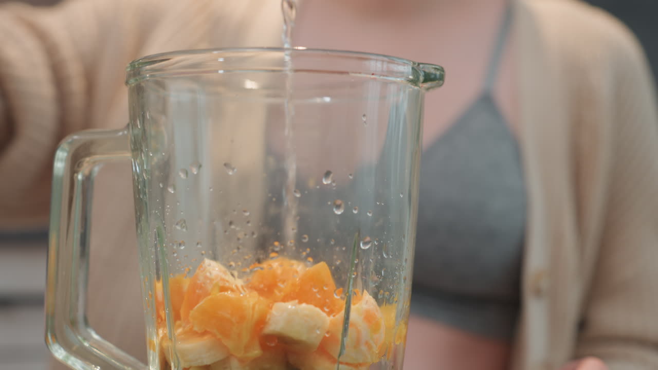 Donna incinta che aggiunge acqua alla frutta nel barattolo del frullatore, spicchi di mandarino e banana visibili, azione di versamento ravvicinata con goccioline sul vetro, focus su texture e idratazione, futura mamma che si prepara