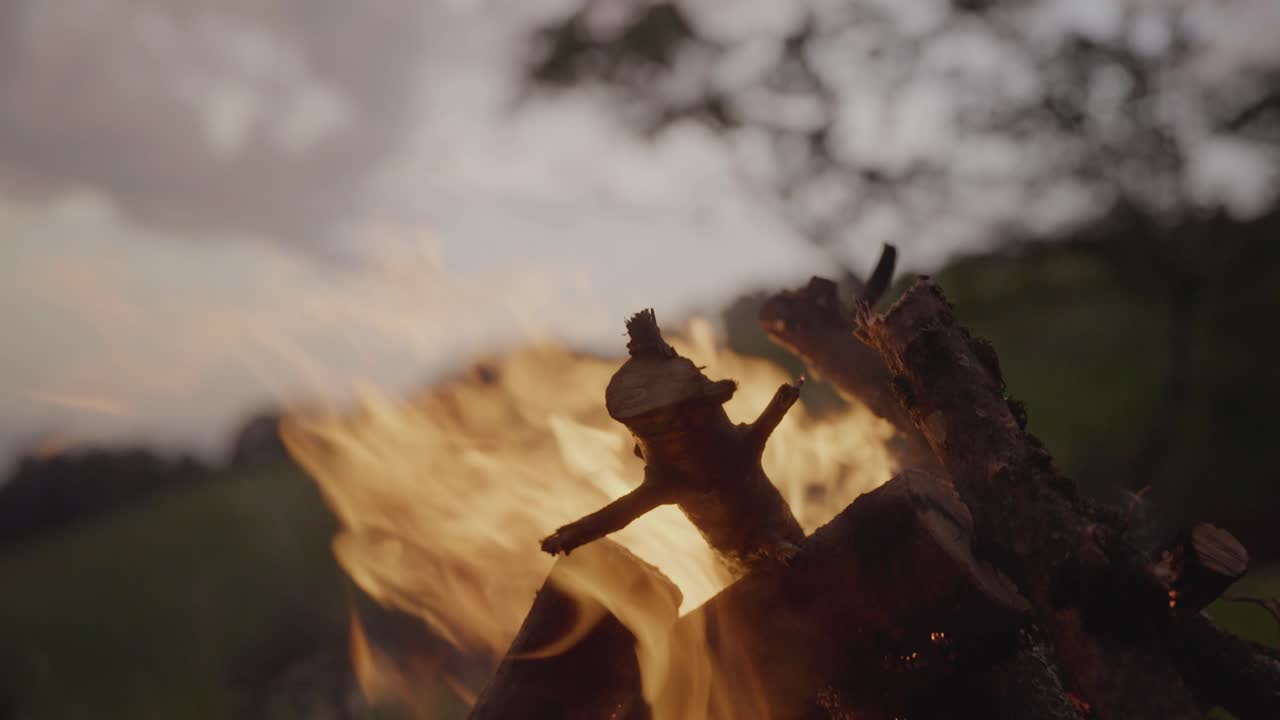 A campfire burns on a grassy hill with a mountain range and lake in the background under a dramatic evening sky. Concept of adventure, nature, solitude, and outdoor lifestyle