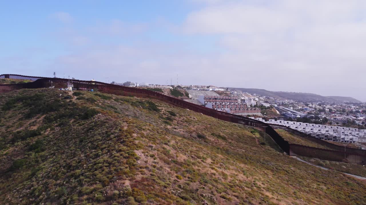 High-elevation drone shot reveals winding border fence snaking over dry hills, separating natural terrain from dense urban sprawl on the horizon.