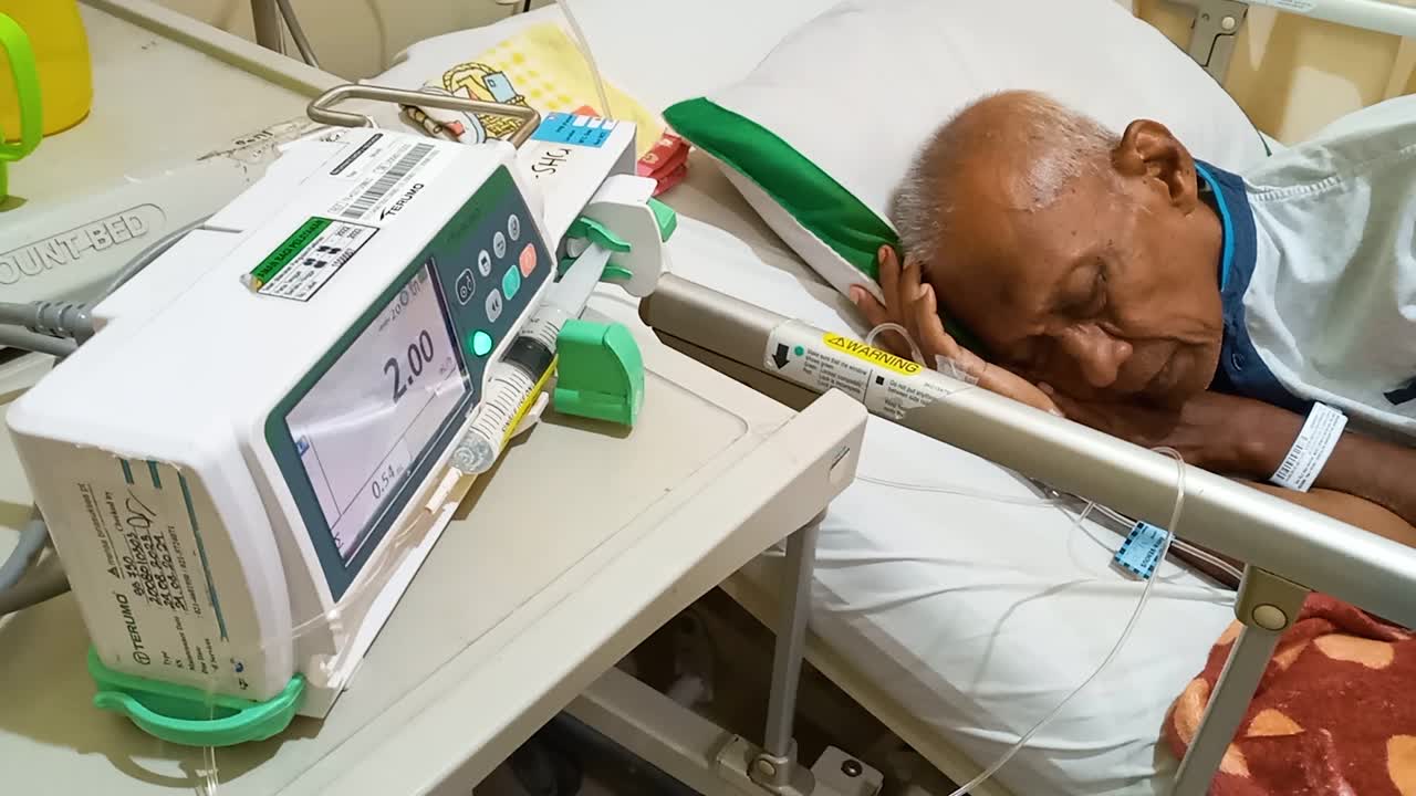 The male patient is asleep on the bed in the hospital, he is using a Syringe pump with a connecting tube and an IV needle in his hand with device light flashes