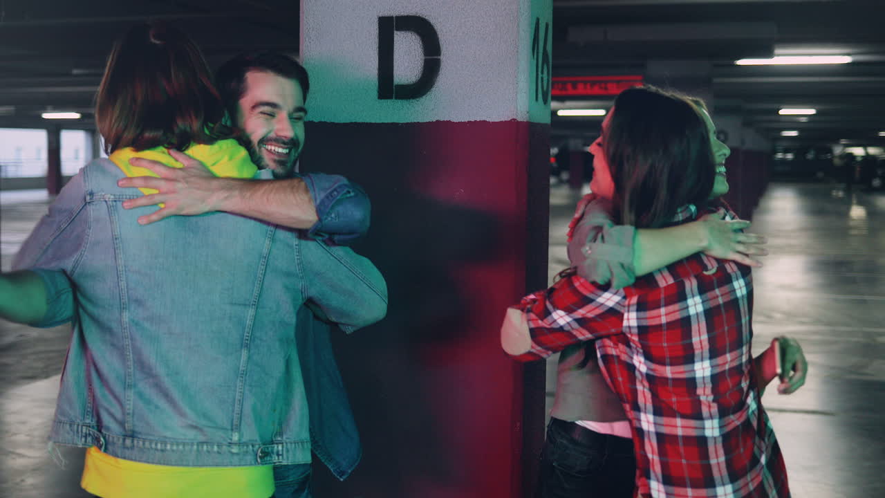 Caucasian young couple standing in a empty parking and using smartphone when their friends man and woman comes up and they greet and hug