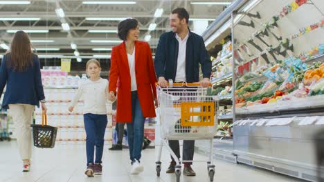 at the supermarket: happy family of three, holding hands, walks through fresh produce section of the store, holding hands. father, mother and daughter having fun time shopping.