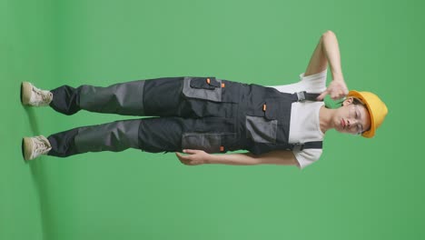 cuerpo lleno de mujer asiática trabajadora con gafas de protección y casco de seguridad mostrando los pulgares hacia abajo gesto a la cámara mientras está de pie en la pantalla verde estudio de fondo