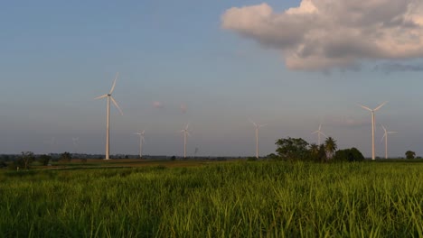 Windkraftanlagen-Schießen-Aus-Einem-Ackerland,-Während-Die-Sonne-Untergeht,-Während-Lastwagen-Vorbeifahren-Und-Staubwolken-Erzeugen,-Saubere-Alternative-Energie-In-Thailand-Und-Auf-Dem-Südostasiatischen-Festland