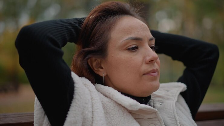 Adult woman leans back on wooden park bench with arms behind head, stretching neck and gathering hair while resting outdoors, soft daylight, serene expression, quiet wellness mood, with woman, adult