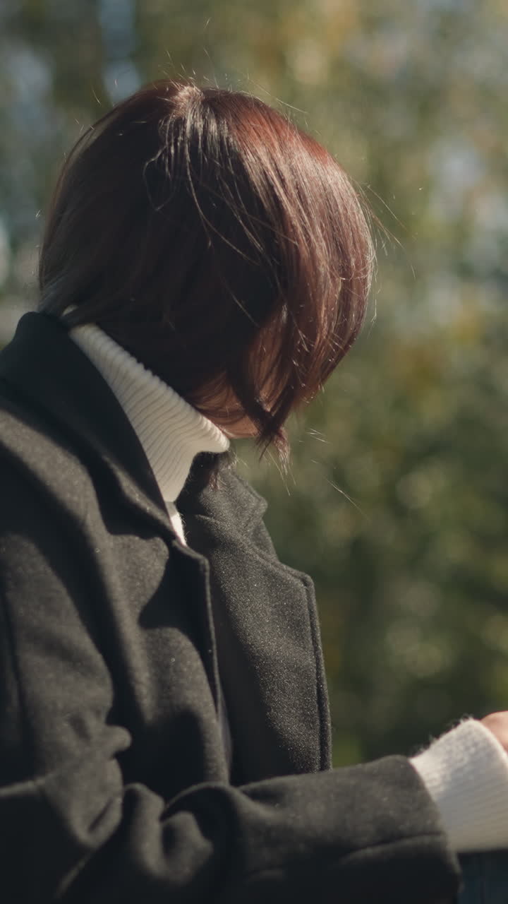 야외에 앉아있는 여자가 사려 깊게 바라보고, 손에 드폰에 집중하고, 손가락에 반지, 은 날 초록색과 나무의 흐릿한 배경과 함께