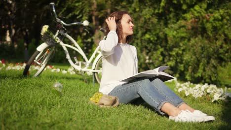 attractive woman in white shirt and blue jeans is reading a book sitting on the grass in park during sunny warm day. her city