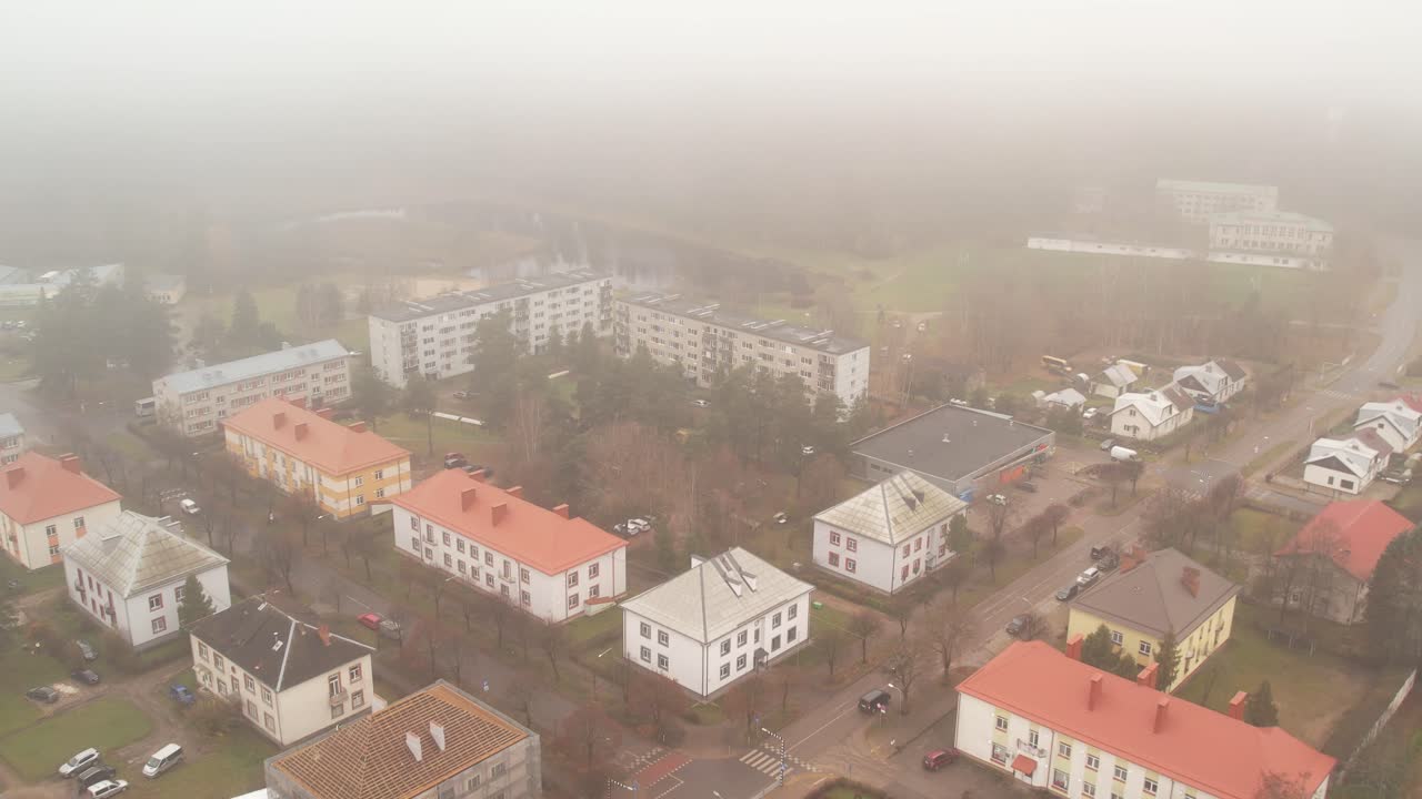 A misty aerial scene of a small town with colorful rooftops, residential buildings, and surrounding trees partially obscured by thick fog