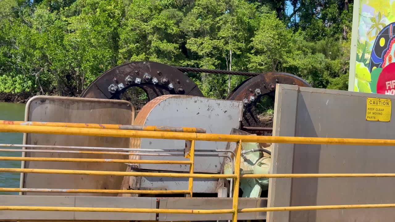 Large industrial cable pulley system in motion on a car ferry, bright daylight, stationary camera, lush riverside vegetation in background, clear mechanical detail