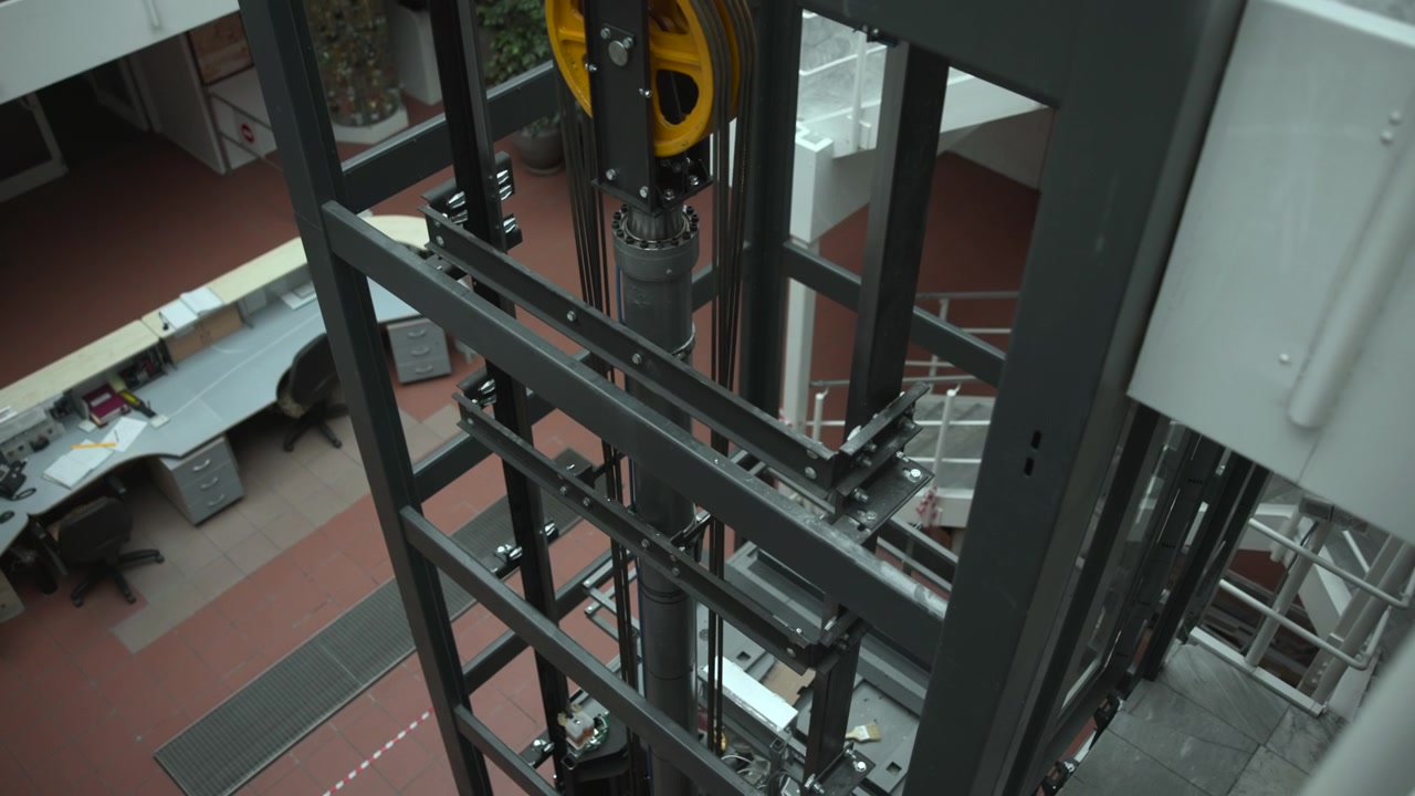 High oblique view into elevator shaft interior showing black steel framework, vertical guide rails, tensioned cables, and central cylinder within industrial maintenance space, with elevator, shaft