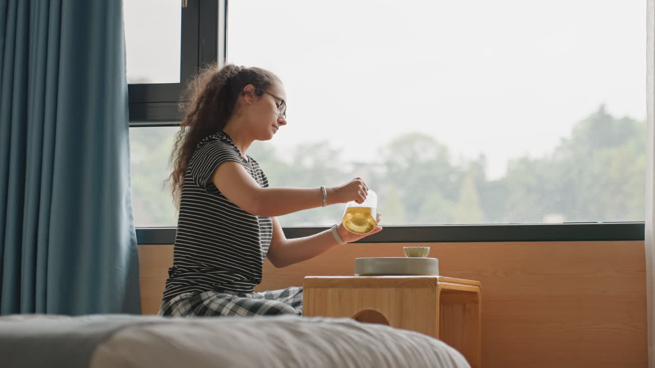 Bambina che fa colazione a bordo letto in hotel, versa succo dalla caraffa nel bicchiere su un piccolo tavolo di legno accanto al letto, vista luminosa dalla finestra con linea di alberi, tende morbide e biancheria da letto dell'hotel in primo piano, scena giocosa.