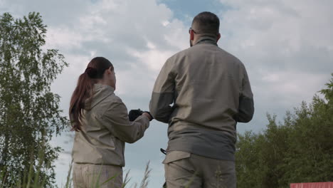 rear view of woman putting on glove while male colleague stands by, both wearing khaki jackets amid grassy field and trees, cloudy sky overhead