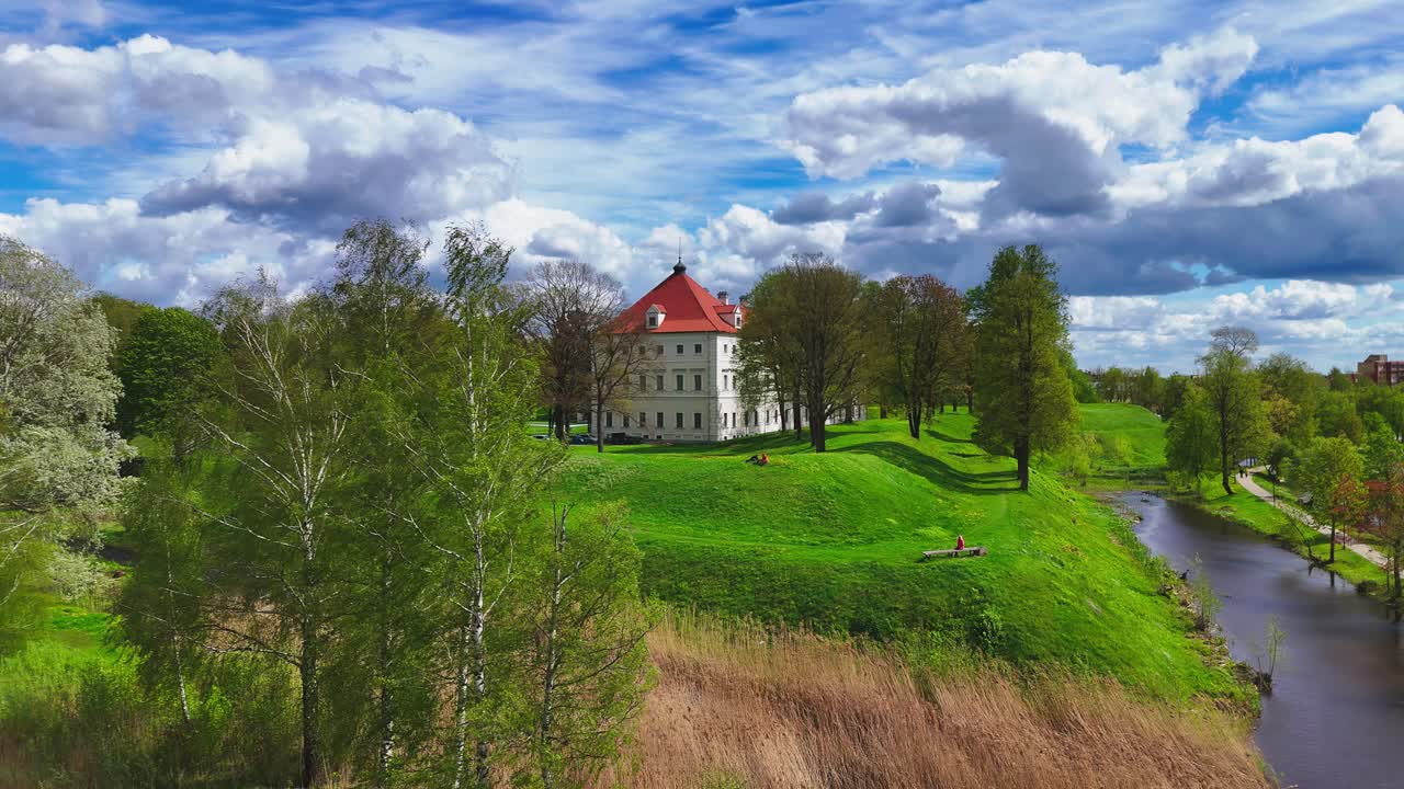 A white manor with a red roof stands atop a green hill bordered by a winding river, spring foliage, and a dramatic sky with scattered clouds overhead.