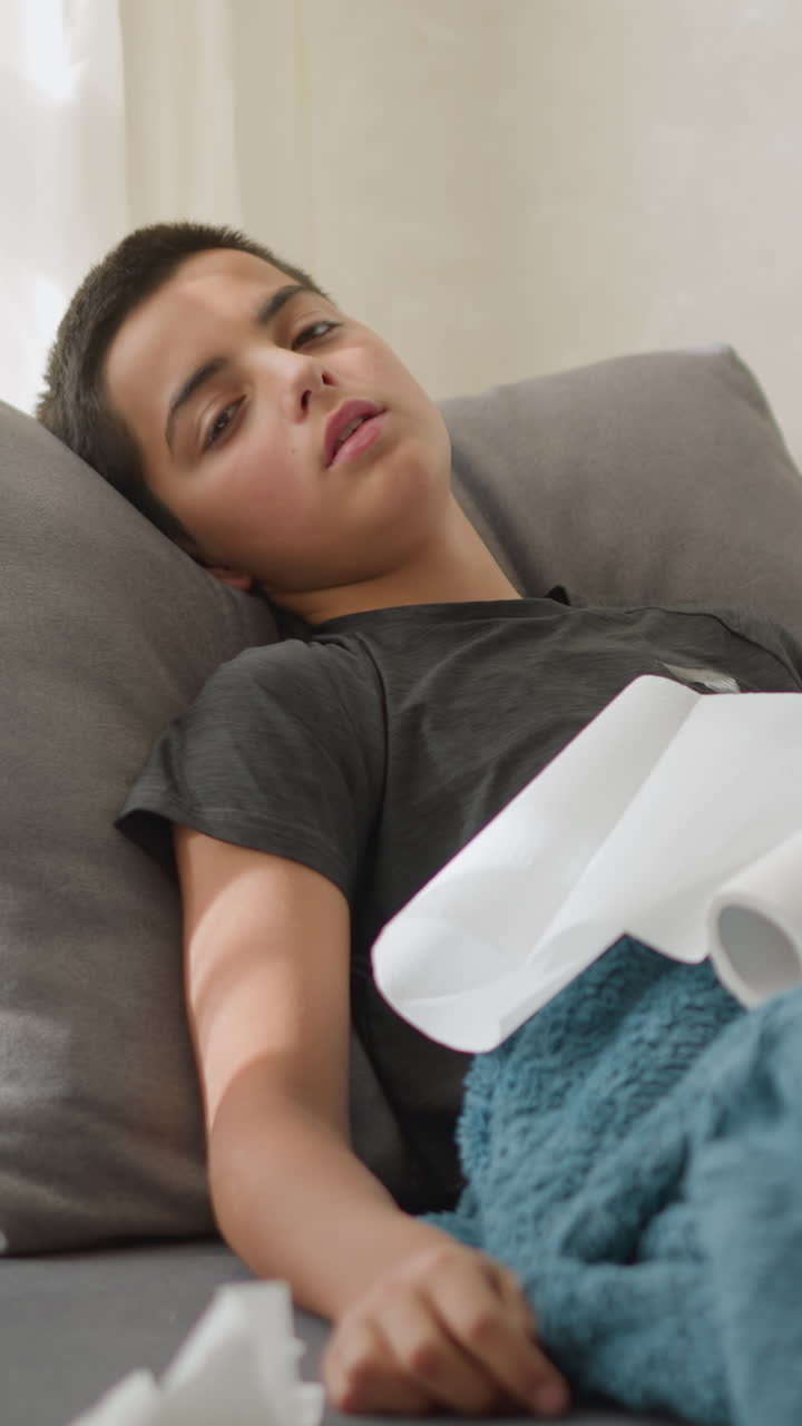 Boy lying down, weak, with tissues scattered around, struggling with illness, covered with blanket, tired and resting at home while fighting flu or cold