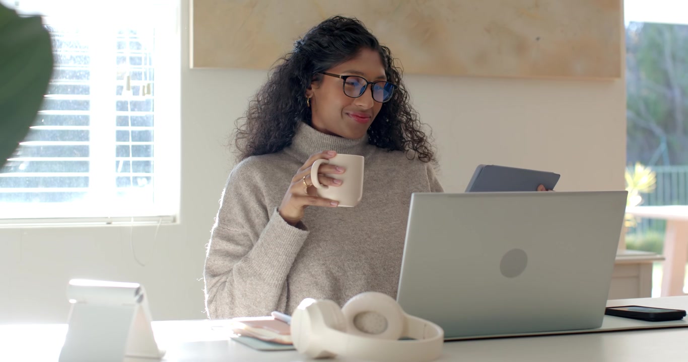 Adult woman wearing grey turtleneck reading tablet alert, holding mug and checking laptop at home