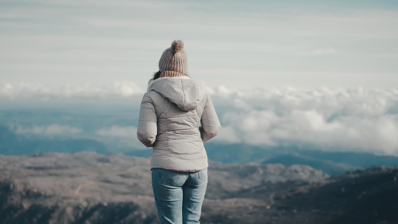 A young Caucasian woman turns her back to the camera and gazes at the mountain landscape