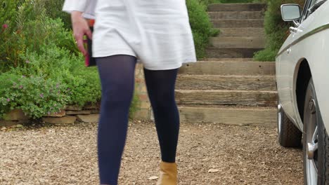 well dressed young lady walks towards camera, down stairs and past vintage car on farm