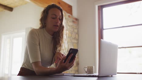 mujer caucásica trabajando desde casa, sentada en un escritorio usando teléfono inteligente y portátil