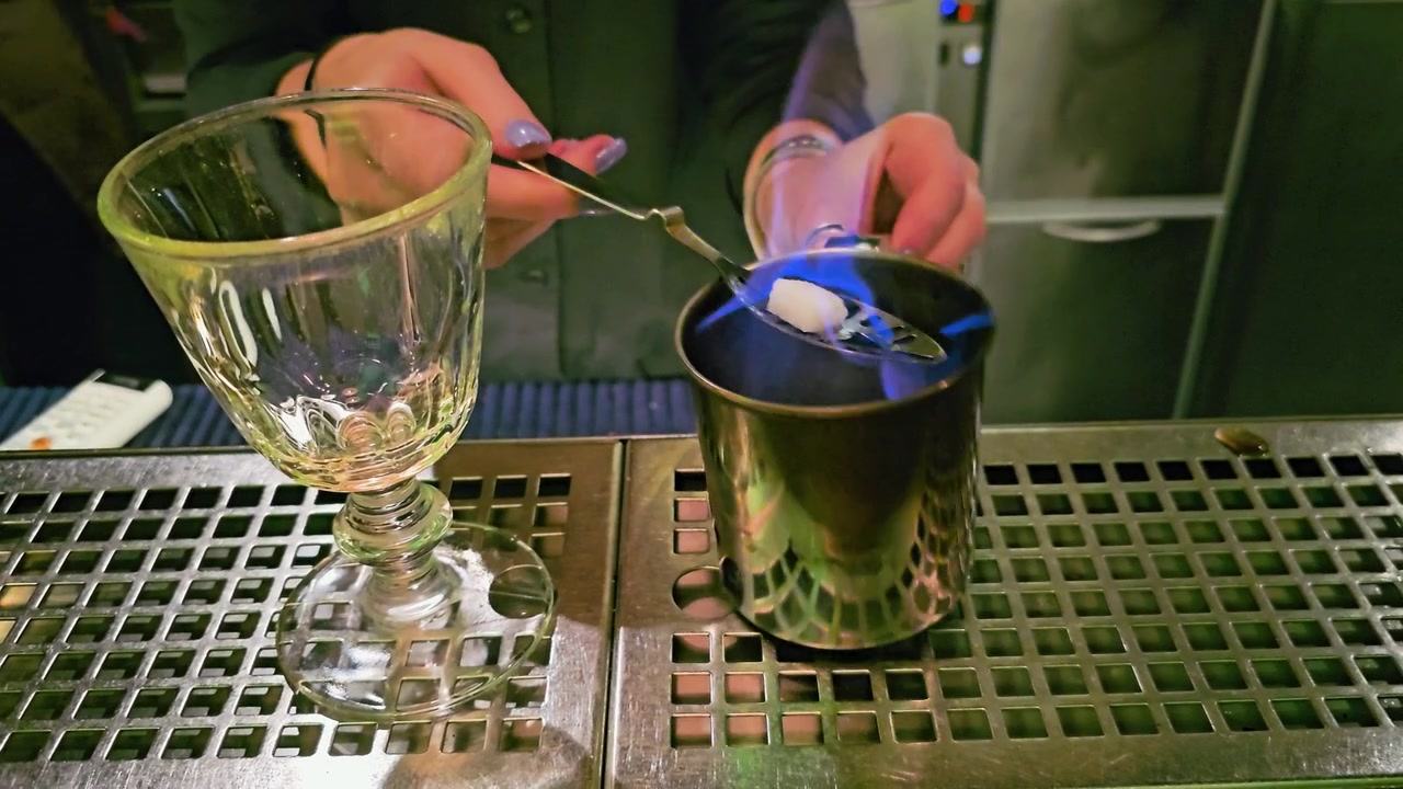 Un barista esegue una tecnica di cocktail infuocata presso una postazione bar in acciaio inossidabile, con fiamme blu brillanti, una zolletta di zucchero, un cucchiaio forato, una tazza di metallo, bicchieri e un'illuminazione verde suggestiva.