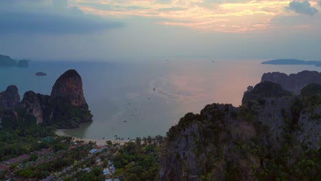 railay beach with traditional longtail boats dotting the andaman sea, surrounded by towering limestone cliffs at sunrise, krabi, thailand. great aerial view flight descending drone