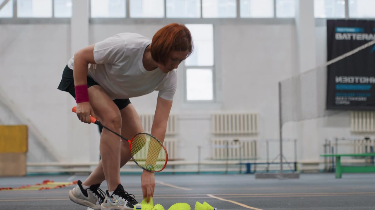 Hvit kvinne serverer badminton i innendørs idrettshall, trenerlærling øver på presisjon, racketer og haug med fjærballer, målte kast og konsentrert holdning, lyse vinduer og metodisk
