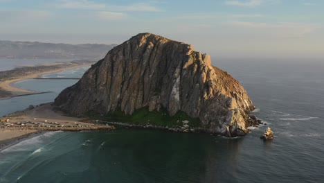 aerial drone circling morro bay rock beach in california usa during sunset as pacific ocean waves crash onto the sandy beach on a summer afternoon