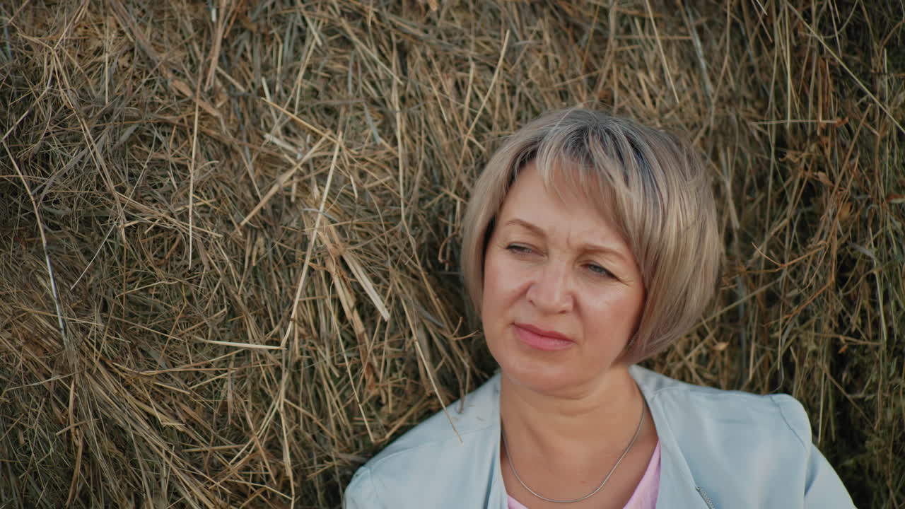 Middle-aged woman rests thoughtfully on hay bale with vast farmland in background, she gazes into the distance, surrounded by rolling fields and a peaceful rural setting
