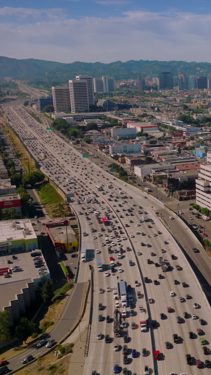 Wide multi-lane freeway full of cars moving along. Flight over sunlit Los Angeles, California, USA. Hazy hills at backdrop. Vertical video