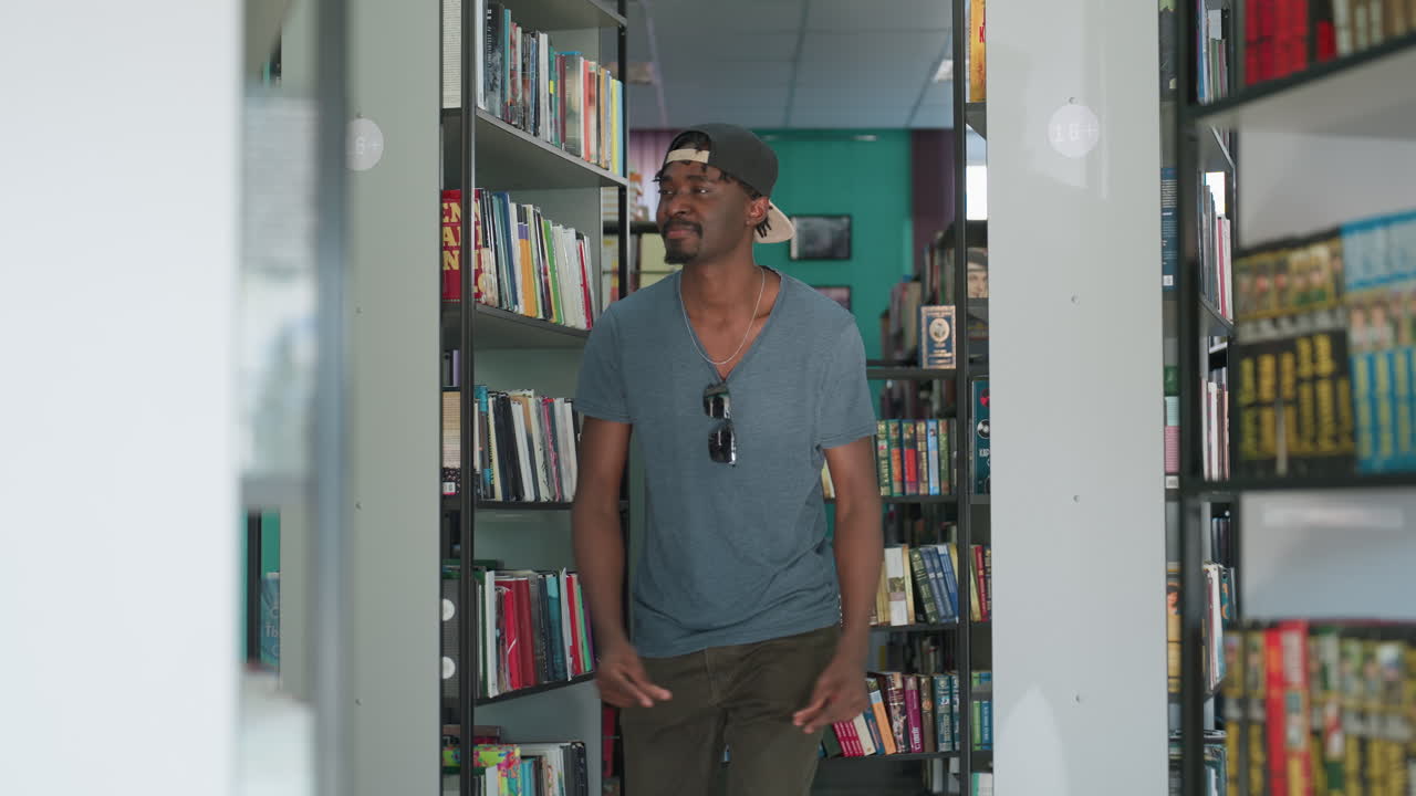 College student wearing backward cap and sunglasses on shirt dancing joyfully between library bookshelves, surrounded by colorful books in bright modern reading space with green walls