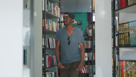 College student wearing backward cap and sunglasses on shirt dancing joyfully between library bookshelves, surrounded by colorful books in bright modern reading space with green walls