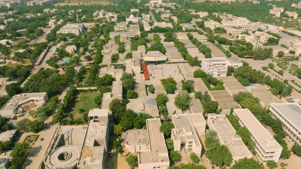 Drone descending and orbiting with a downward tilt, showcasing the green vegetation, trees, and architecture of NED University campus during an intense sunny day